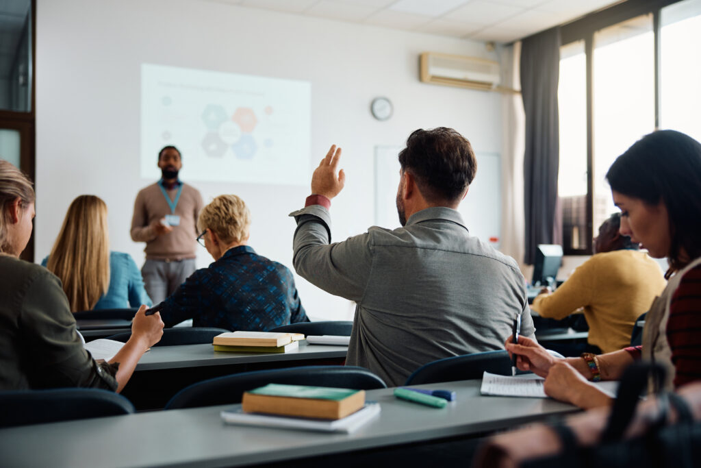 back view of older student raising his hand to answer teacher's question during education training class.